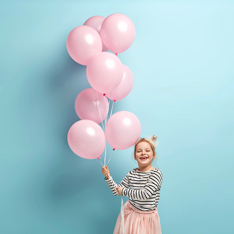 Positive small kid dressed in festive clothes, carries air balloons, celebrates holiday, has broad smile, stands against blue background, being in high spirit. Children and celebration concept. Kinderyoga Venray